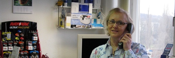 Frau im Büro telefoniert, im Hintergrund Regale mit Hörgerätebatterien und Zubehör.